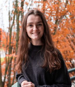 Female math student leans on a railing with fall trees in the background