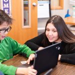 Elementary student typing on class computer while a young student educator assists them