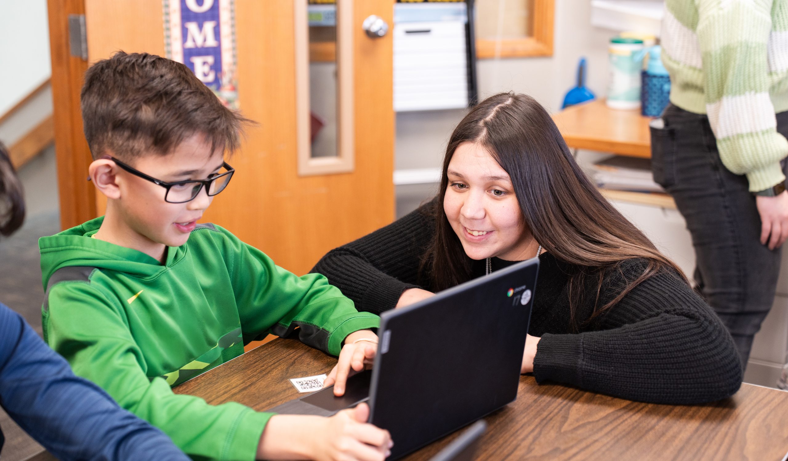 Elementary student typing on class computer while a young student educator assists them