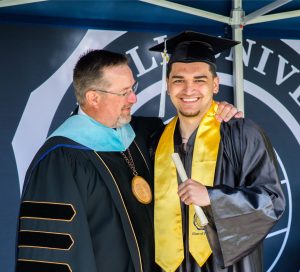 Male student in graduation cap and gown holding their degree with Bushnell University President Dr. Joseph Womack