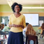 A young teacher smiles with an elementary classroom in the background
