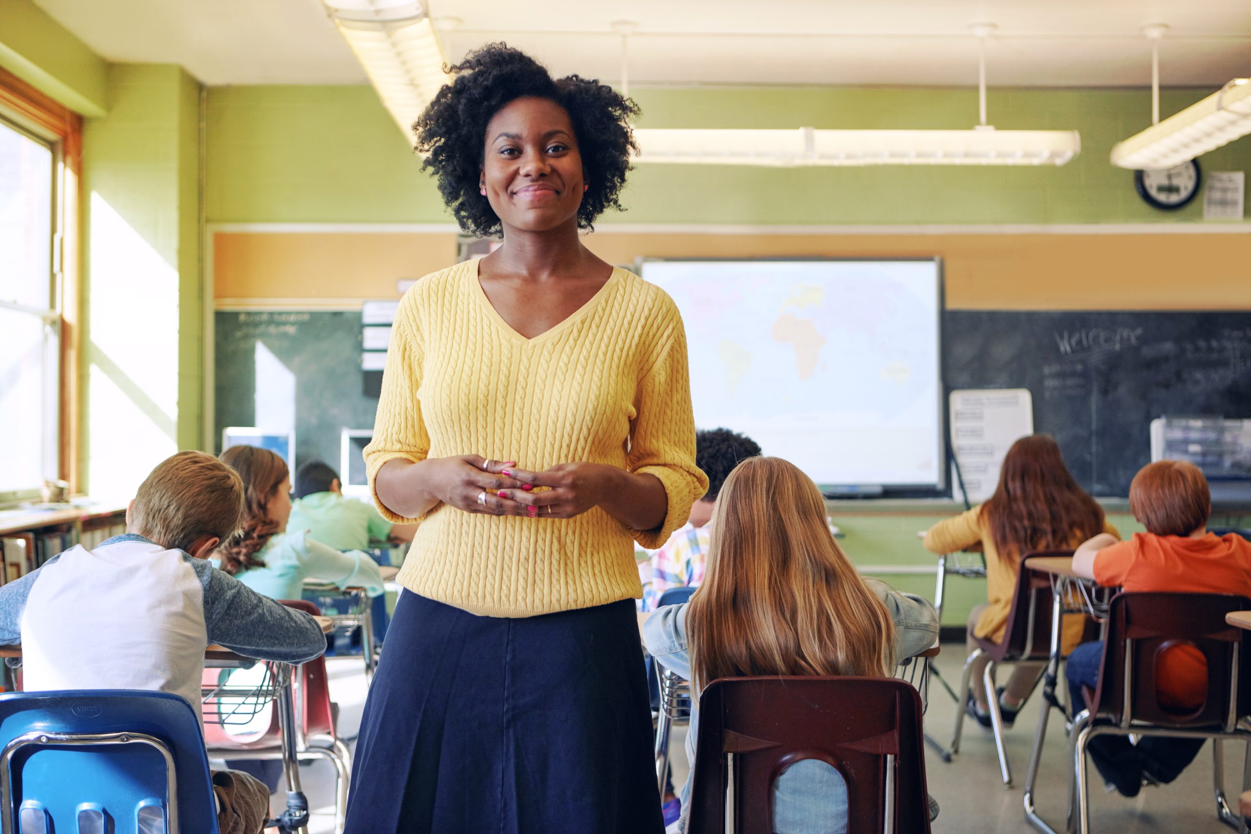 A young teacher smiles with an elementary classroom in the background