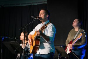 Music program director leading Bushnell worship band in song while holding guitar