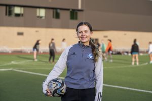 A young soccer coach holds a soccer ball as athletes practice behind her