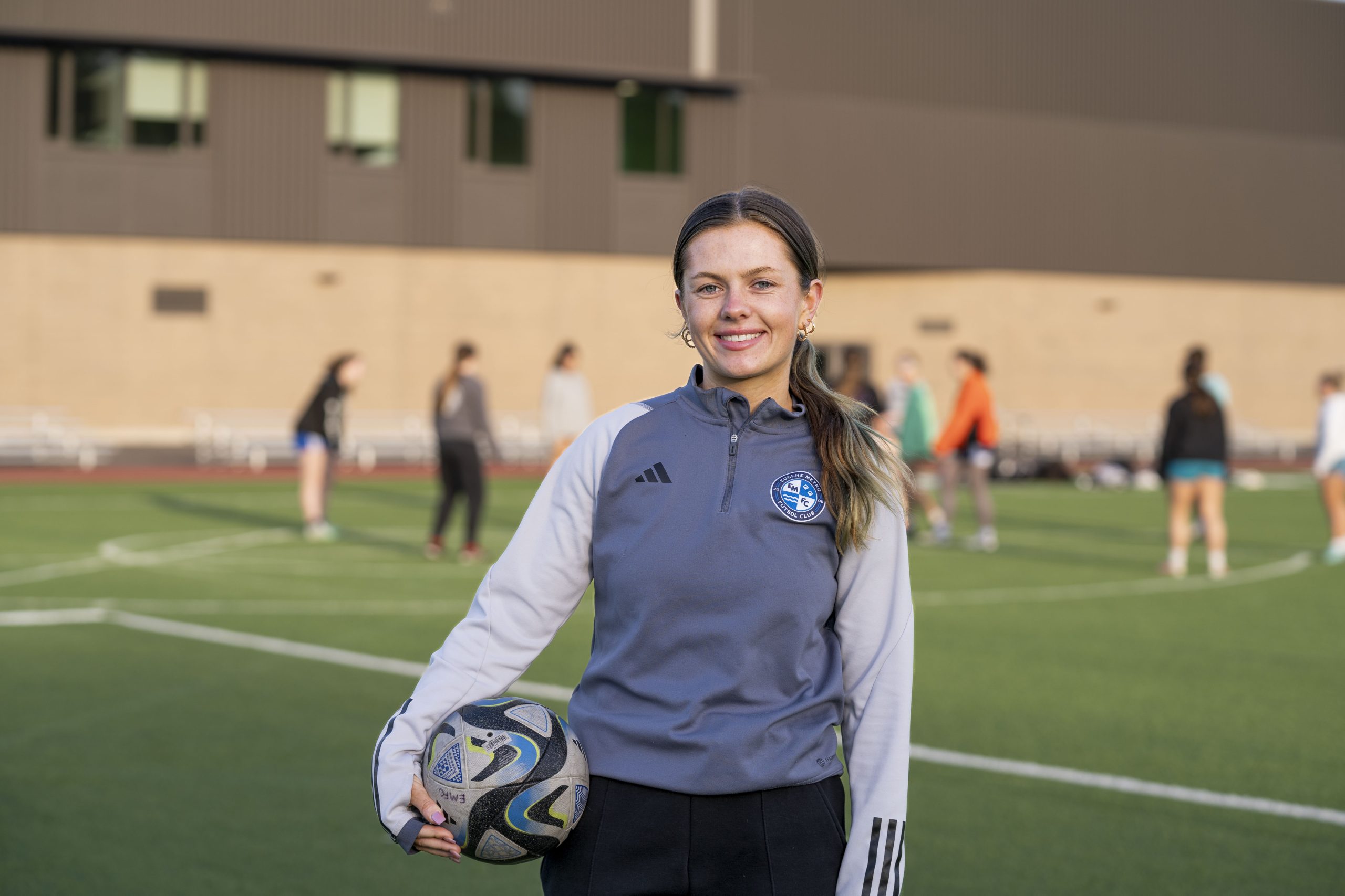 A young soccer coach holds a soccer ball as athletes practice behind her