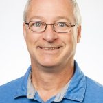 Professional headshot of Bryan Olmstead wearing a blue collared shirt and glasses.