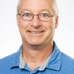 Professional headshot of Bryan Olmstead wearing a blue collared shirt and glasses.