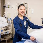 Bushnell University nursing student prepares a patient bed in a clinical simulation lab while wearing scrubs and a stethoscope.