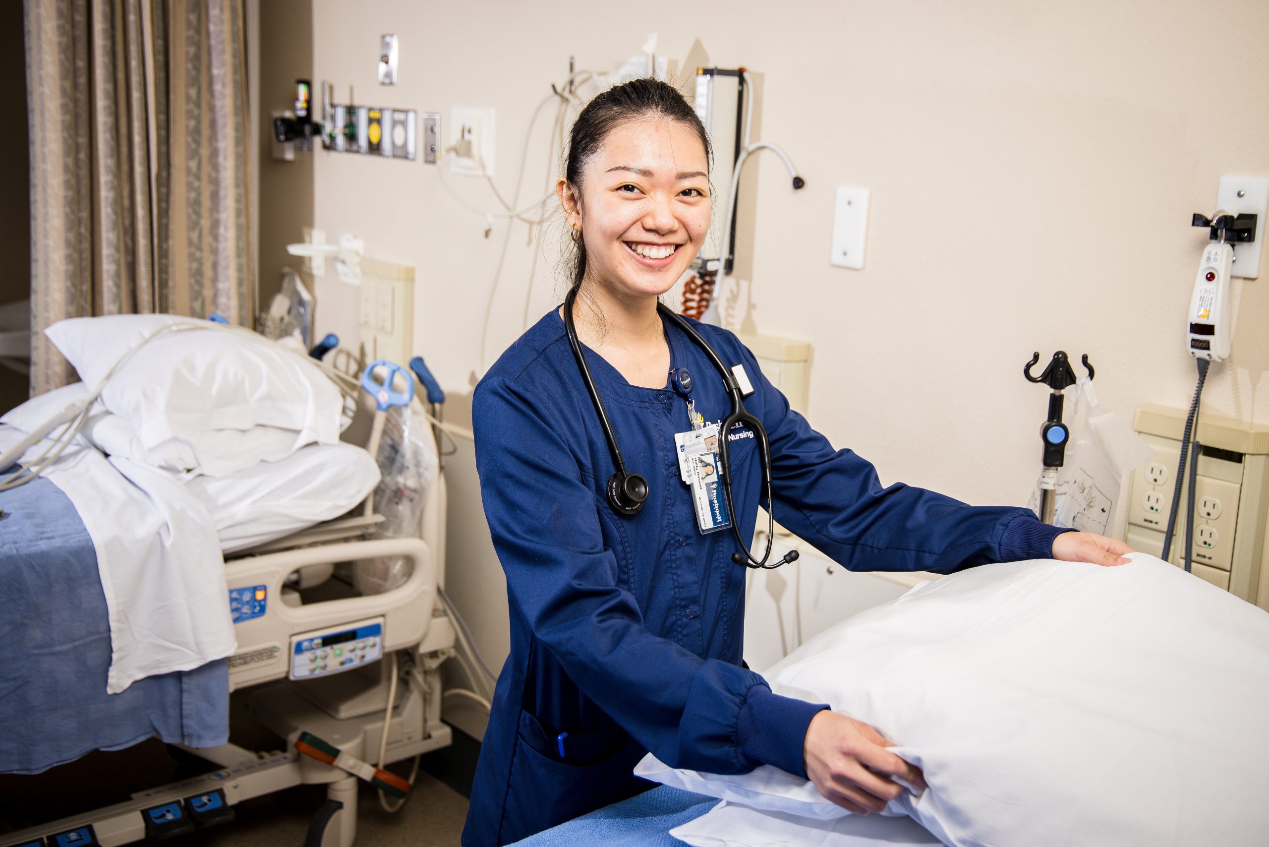 Bushnell University nursing student prepares a patient bed in a clinical simulation lab while wearing scrubs and a stethoscope.