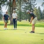 A group of golfers watch one member putt a golf ball on a bright green course during a sunny day.