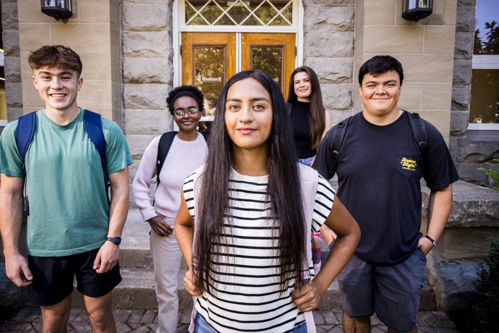 Five Bushnell University students stand together outside a campus building, smiling and carrying backpacks between classes.