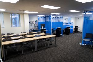 Nursing learning laboratory with classroom seating, patient simulation stations, hospital equipment, and privacy curtains.