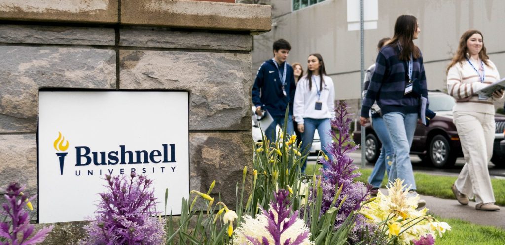 Prospective students walk past a Bushnell University campus sign and landscaped garden during a campus visit.