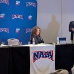 Bushnell University representatives participate in a panel discussion at the NAIA National Convention, with a presenter speaking from the podium and attendees seated at the front table.