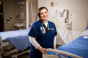 Bushnell University nursing student wearing scrubs and a stethoscope standing in a clinical simulation hospital room.