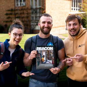Three Bushnell University students smile on campus while holding and pointing to a copy of The Beacon, the university's student publication.