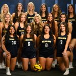 Members of the Bushnell University women's beach volleyball team pose for an official team portrait in Beacons uniforms with a volleyball displayed in front.