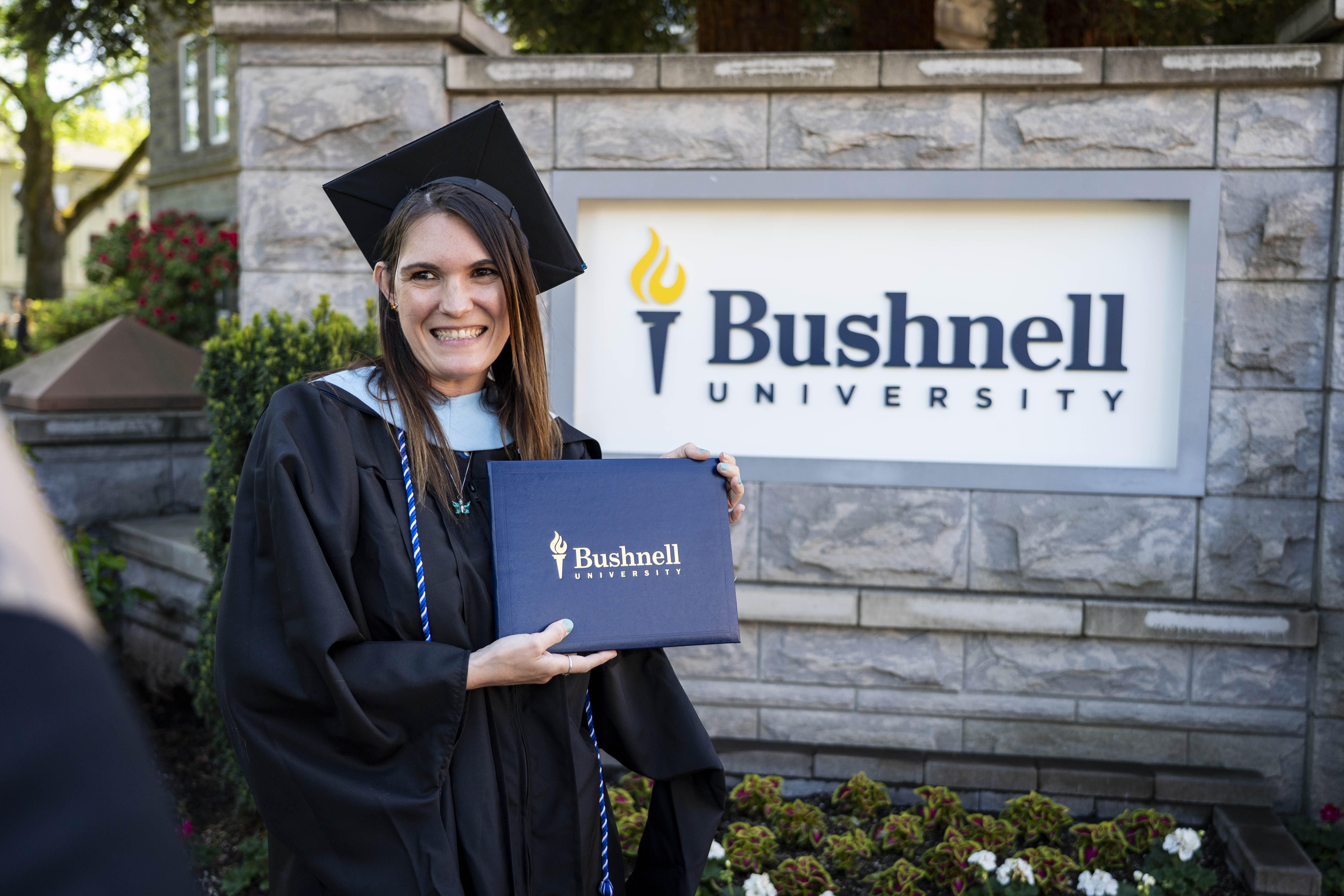 A smiling graduate student in graduation robes and cap holds a Bushnell diploma in front of the university monument sign.