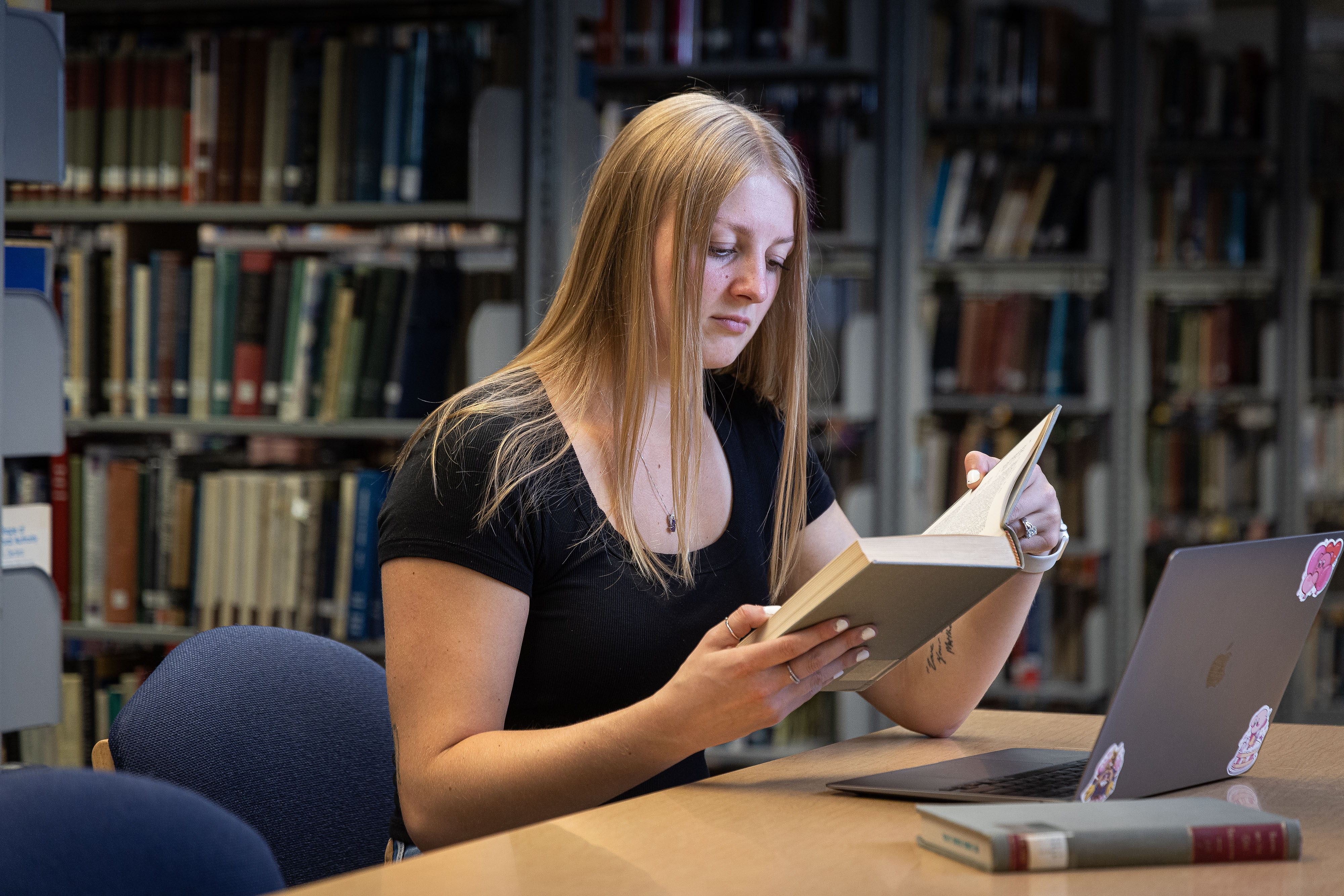 A student reviews a book with their compute in a library setting.