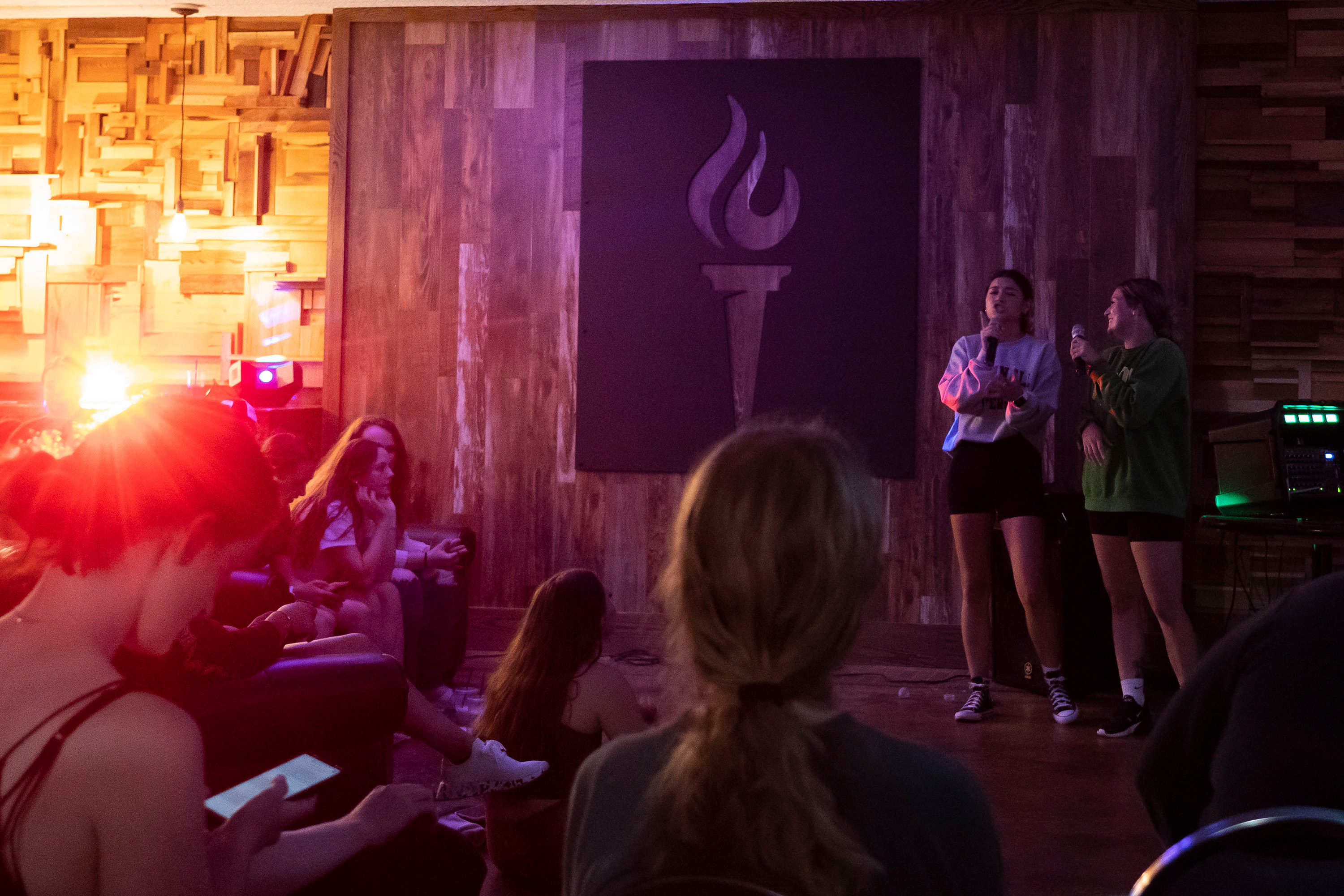 Two students having fun performing karaoke to a crowd with colorful lights in a recreation room.