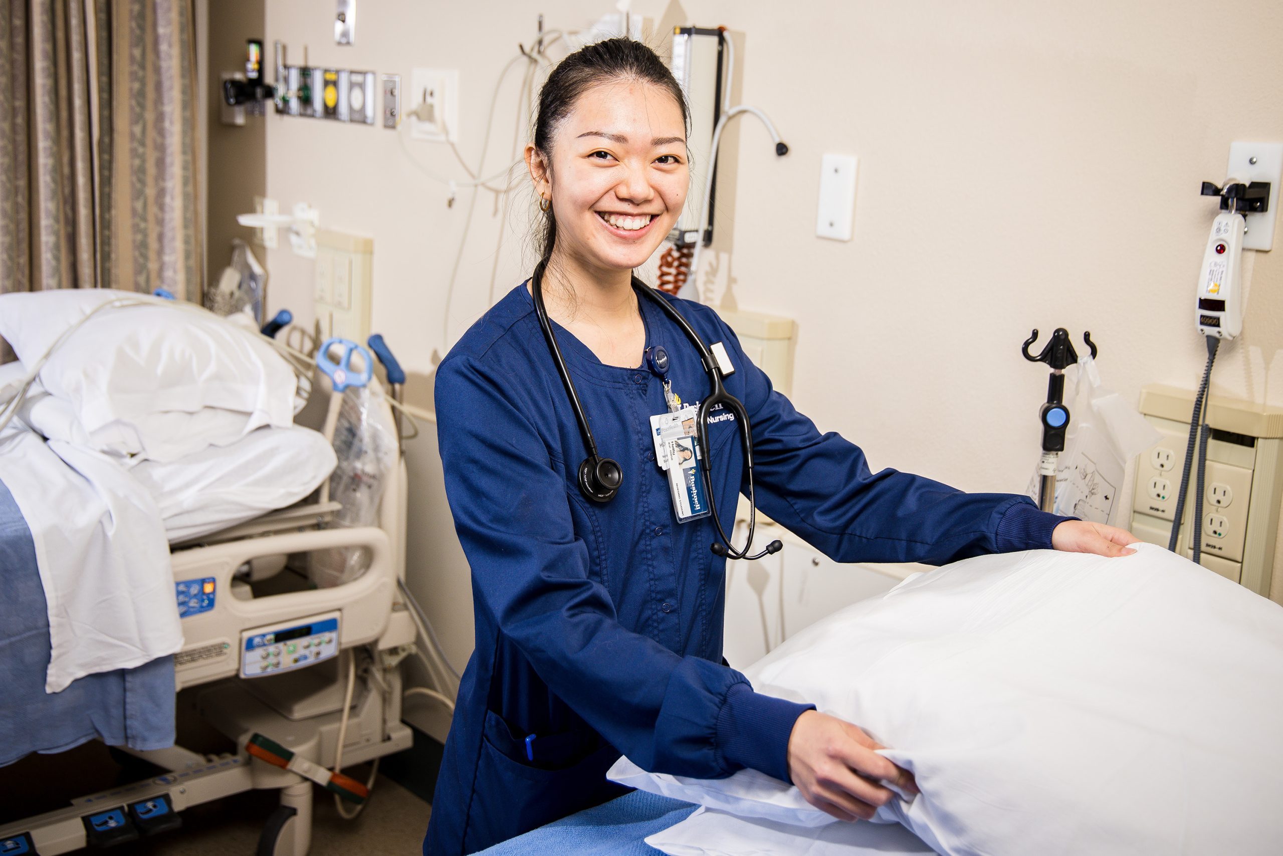 A Bushnell University ABSN student in navy blue scrubs and stethoscope prepares a patient bed.