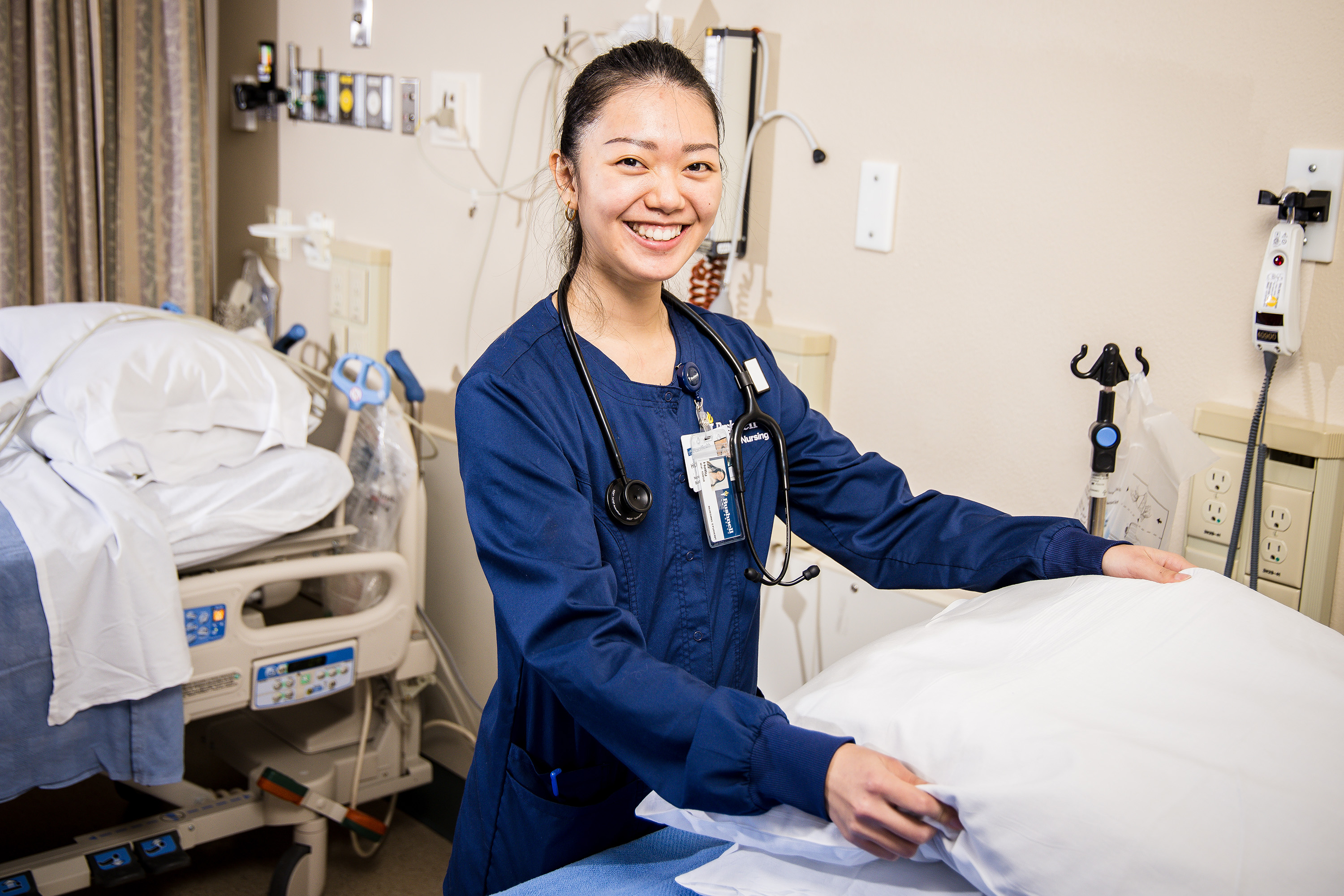 A Bushnell University ABSN student in navy blue scrubs and stethoscope prepares a patient bed.
