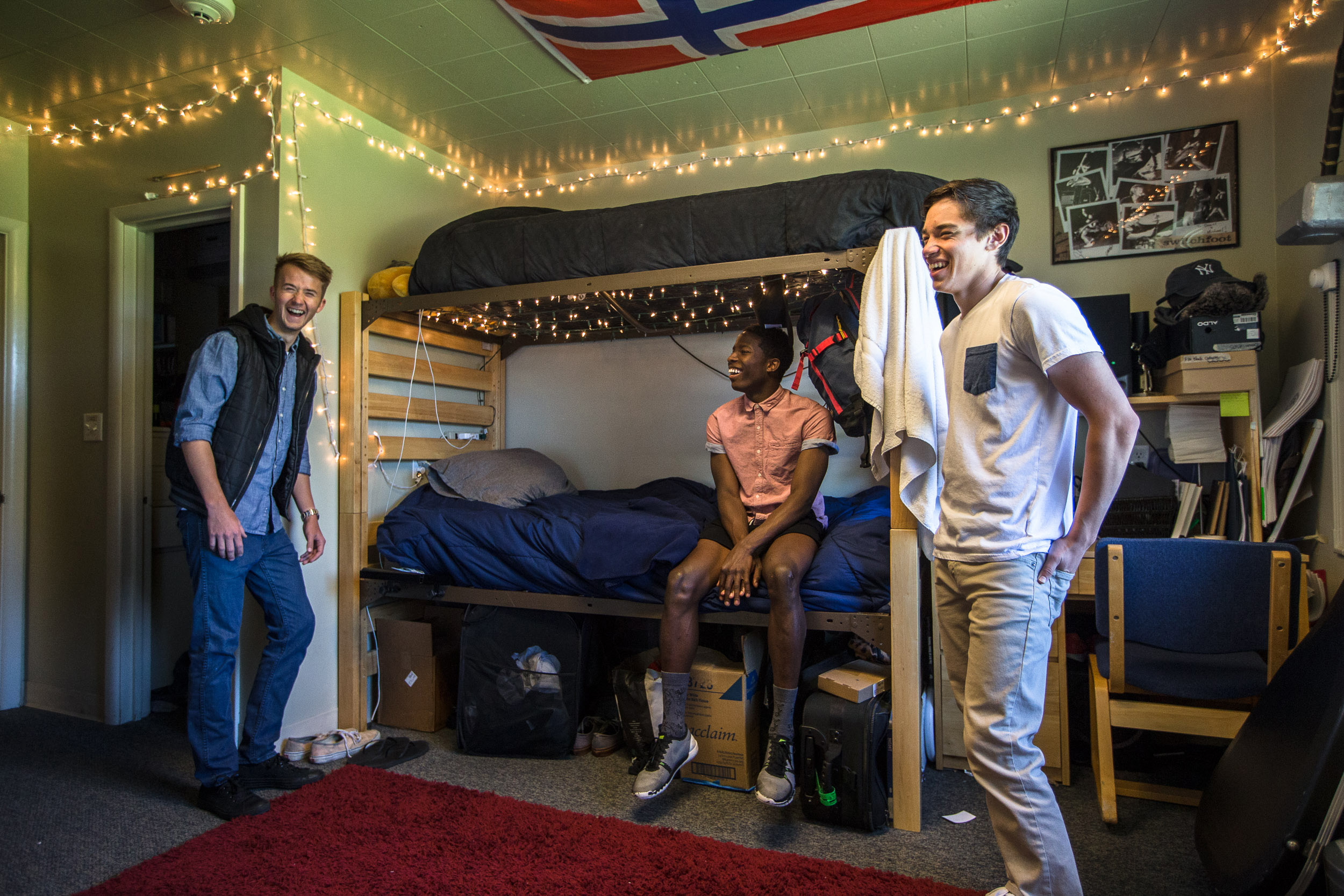 A group of boys in a decorated dorm room with a bunk bed and desks laugh.