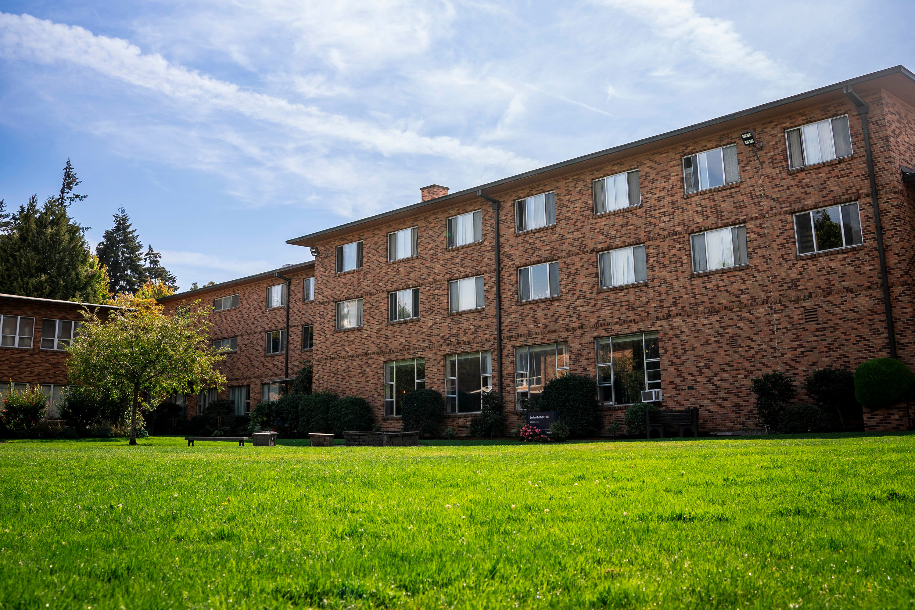 An exterior photo of a orange and brown brick three floor student dorm building with a grassy quad field in front of it.