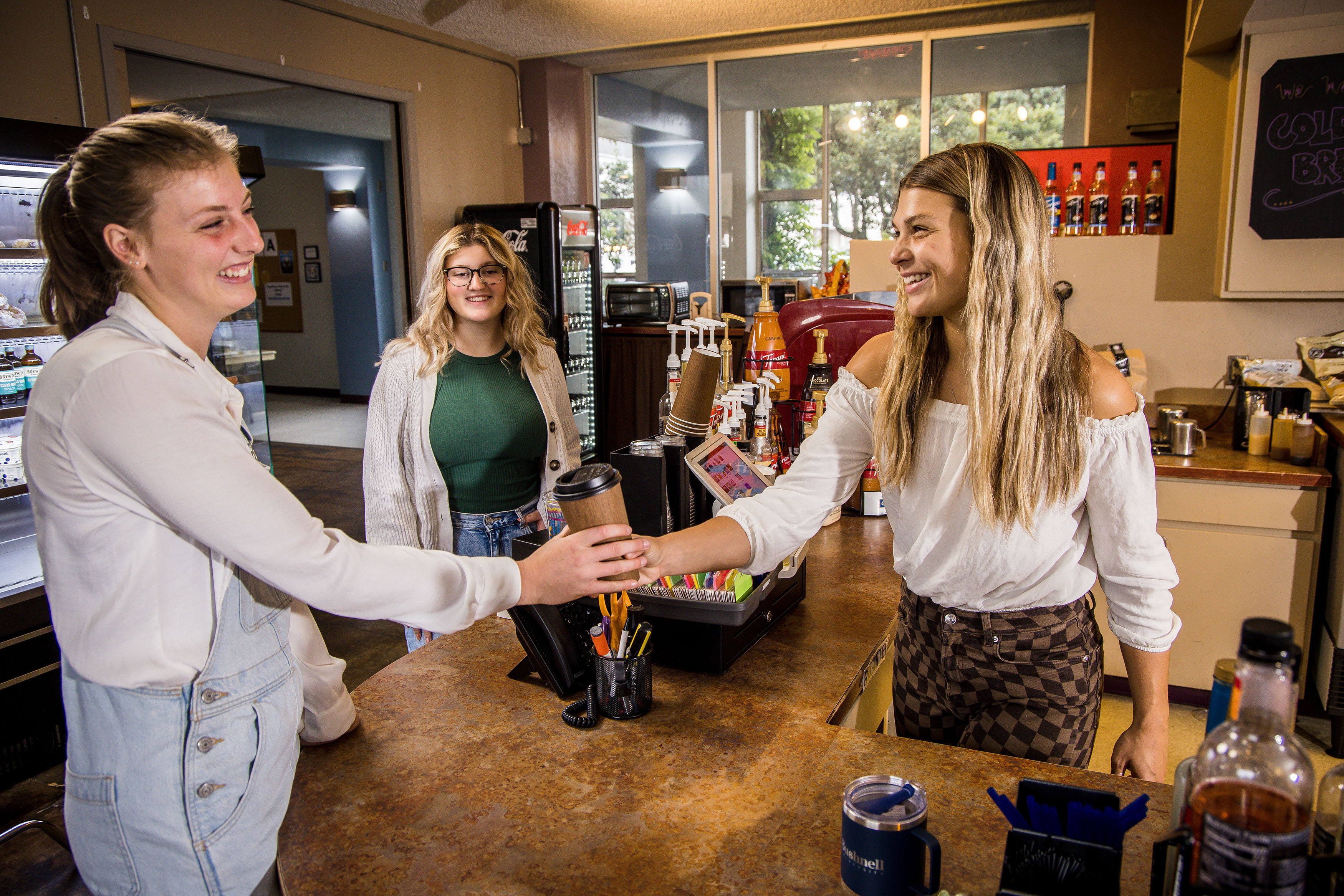 A student barista hands off a prepared coffee cup to another student from behind a cafe counter.