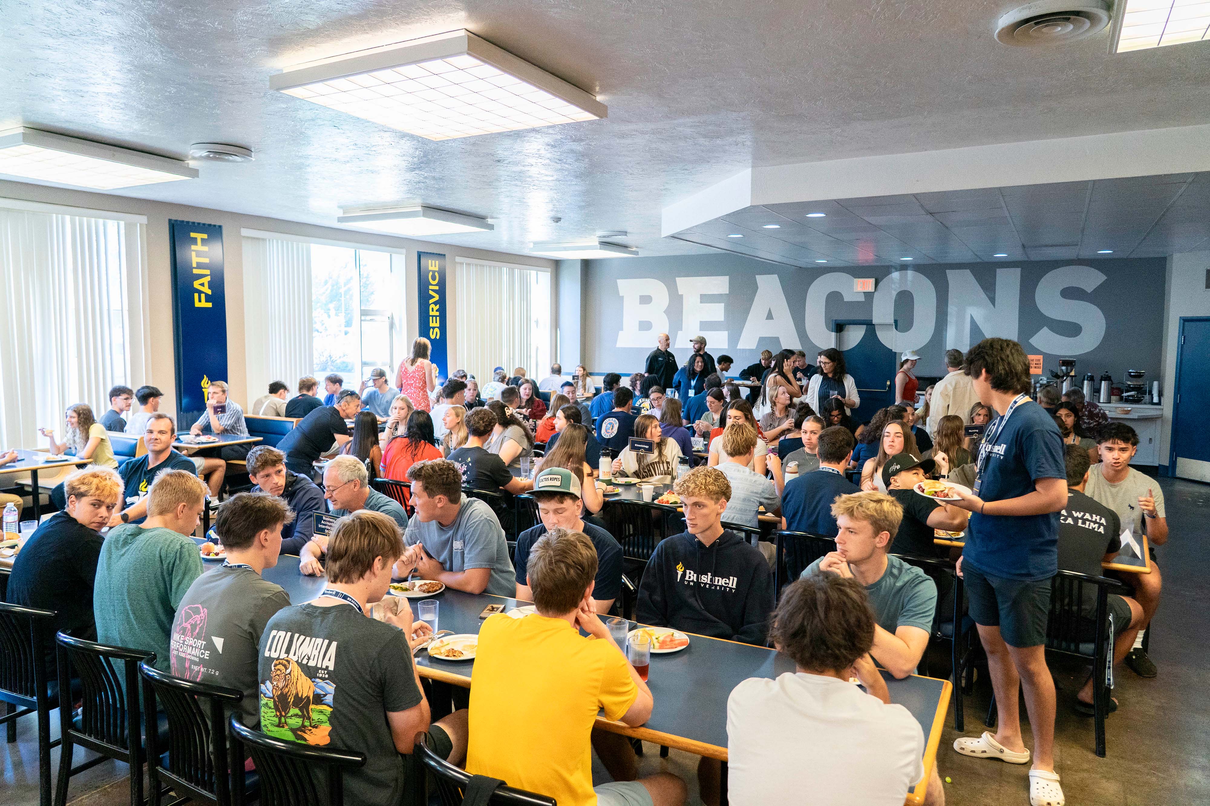 Bushnell University cafeteria with multiple long tables filled with students eating food, booths along a wall with large windows, and the word "Beacons" in big bold letters on another wall.