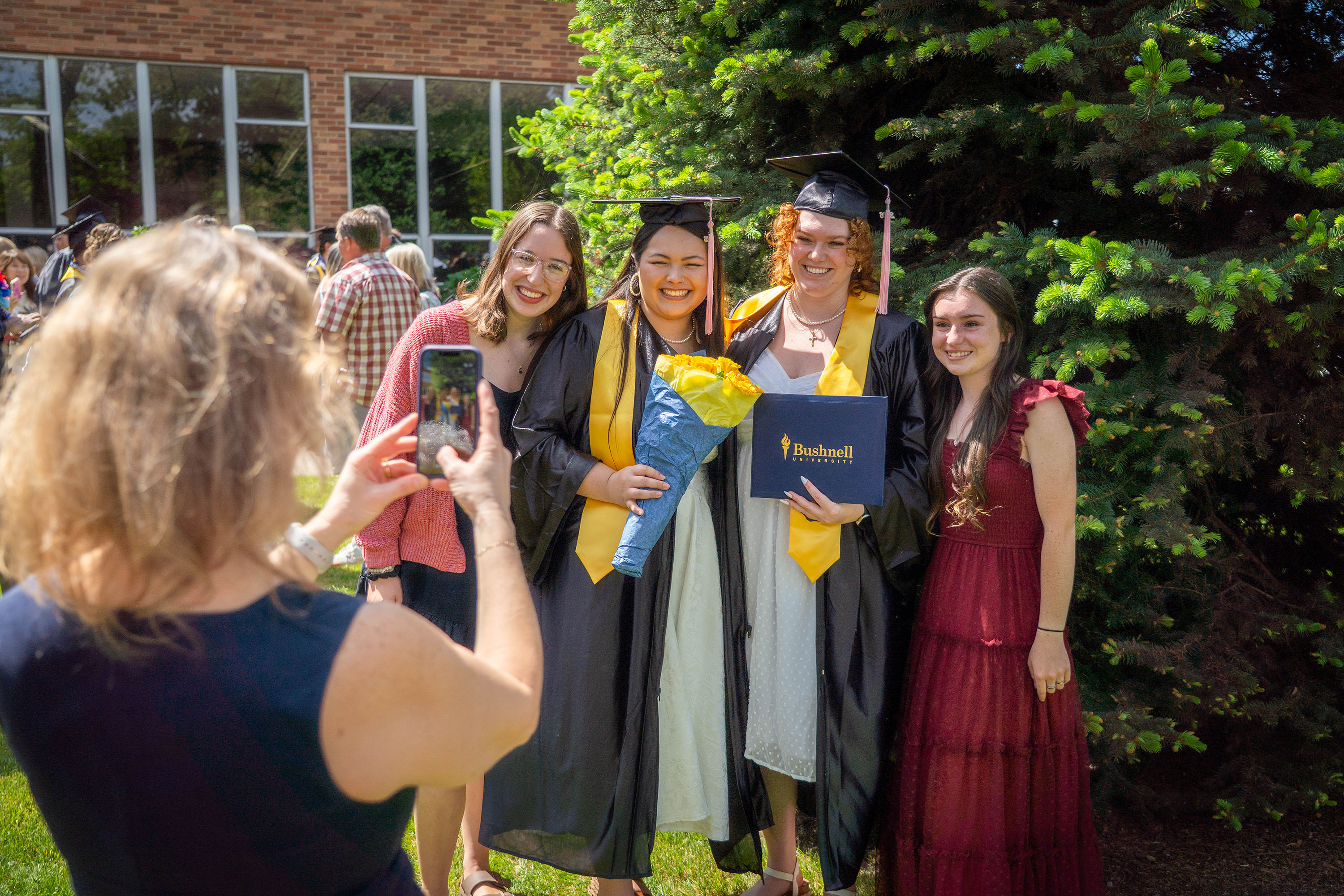 A group of friends take pictures together after a Bushnell University Commencement ceremony holding flowers and diplomas.