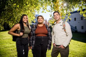 Three Bushnell University students stand together on campus beneath a tree.