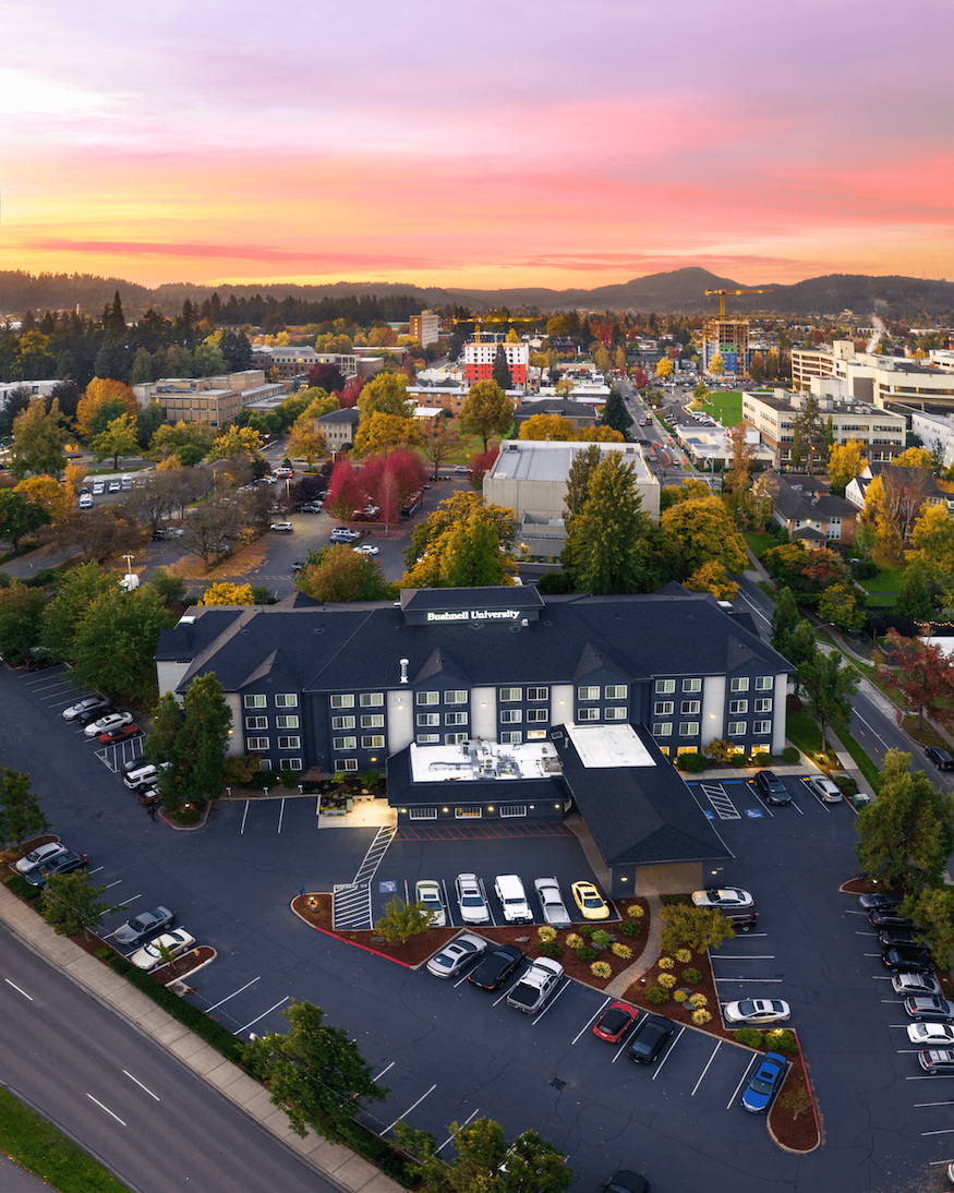 Aerial view of Bushnell University's campus in Eugene, Oregon, at sunrise with colorful autumn trees and the city skyline in the background.