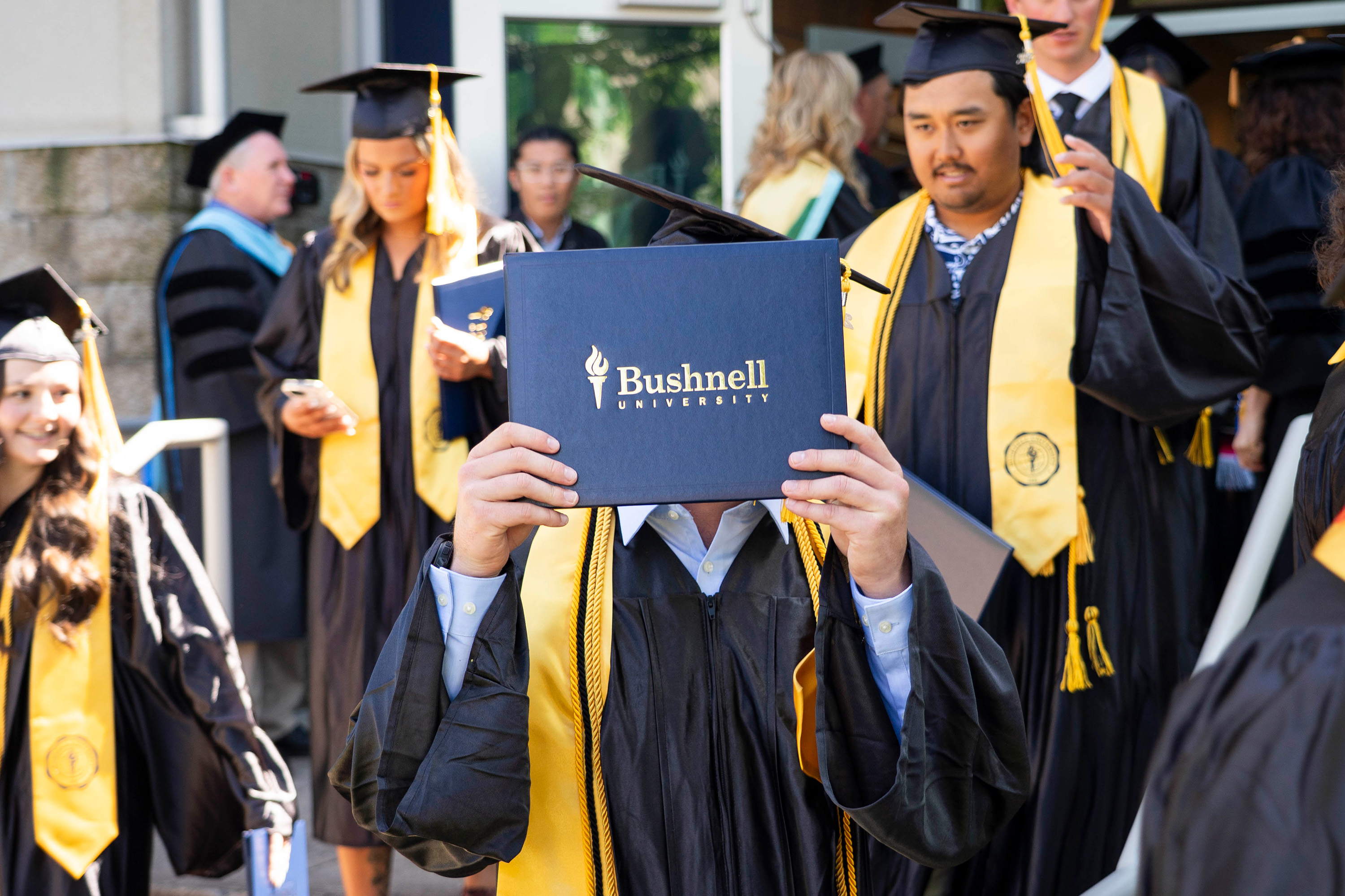 A student holds a Bushnell University diploma in front of of their face while being surrounded by graduating students.