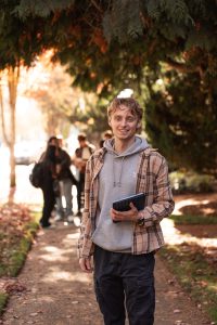 Bushnell University student walking along a tree-lined campus path while carrying a tablet.