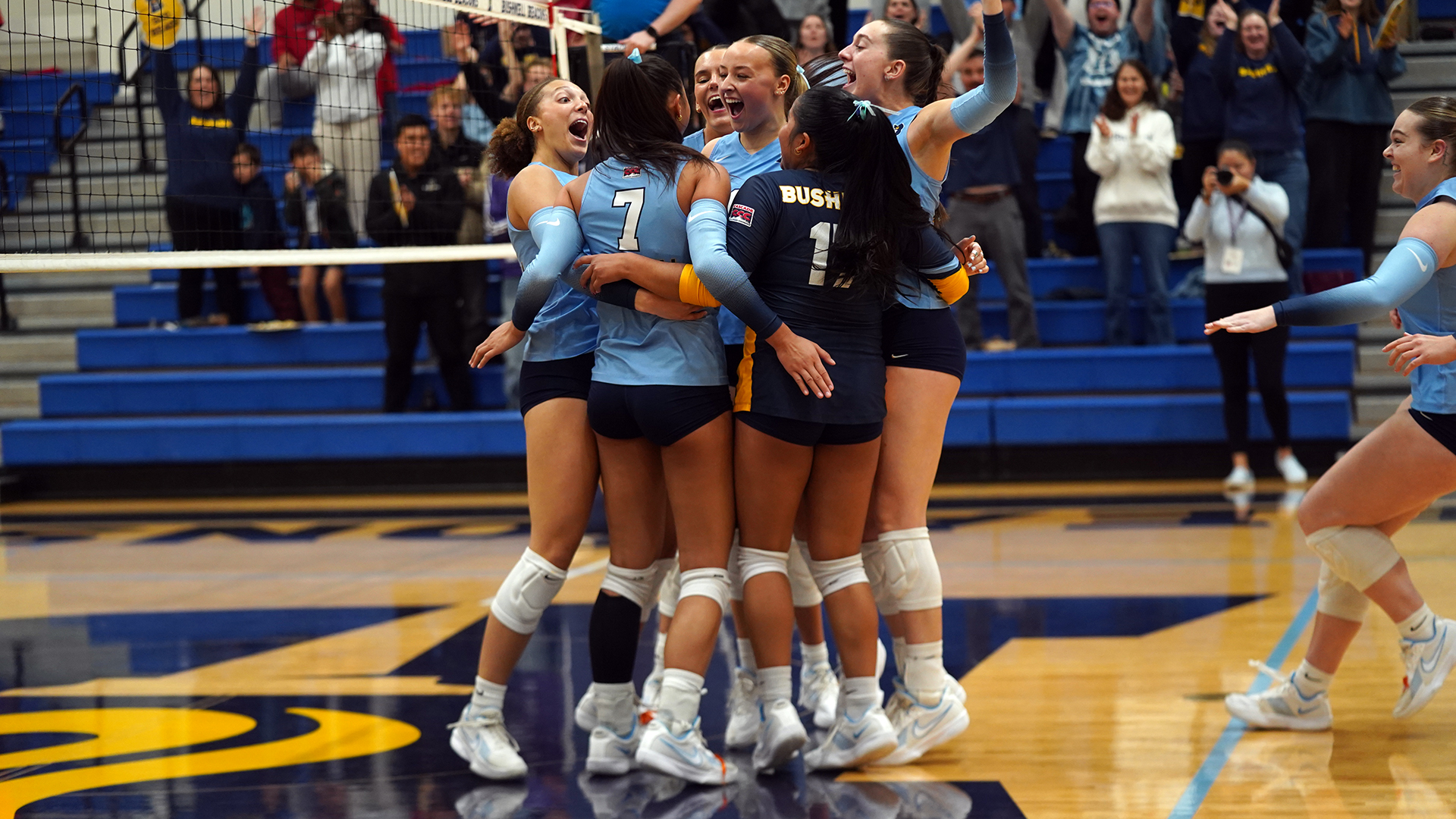 Bushnell University women's volleyball players celebrate together on the court after winning a point as fans cheer in the background.