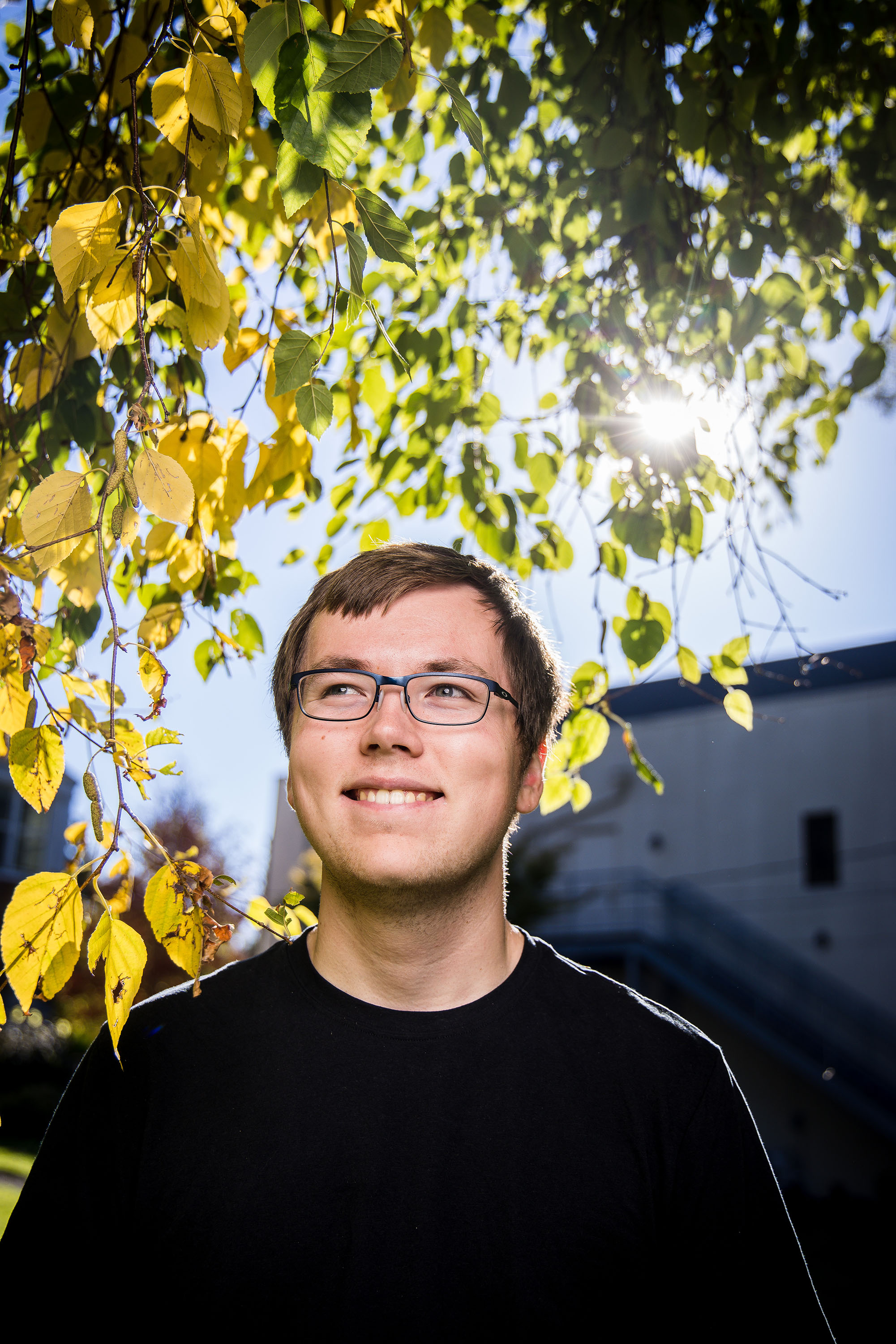 A student with glasses and a black shirt looks up and smiles with sun and leafy branches behind them.
