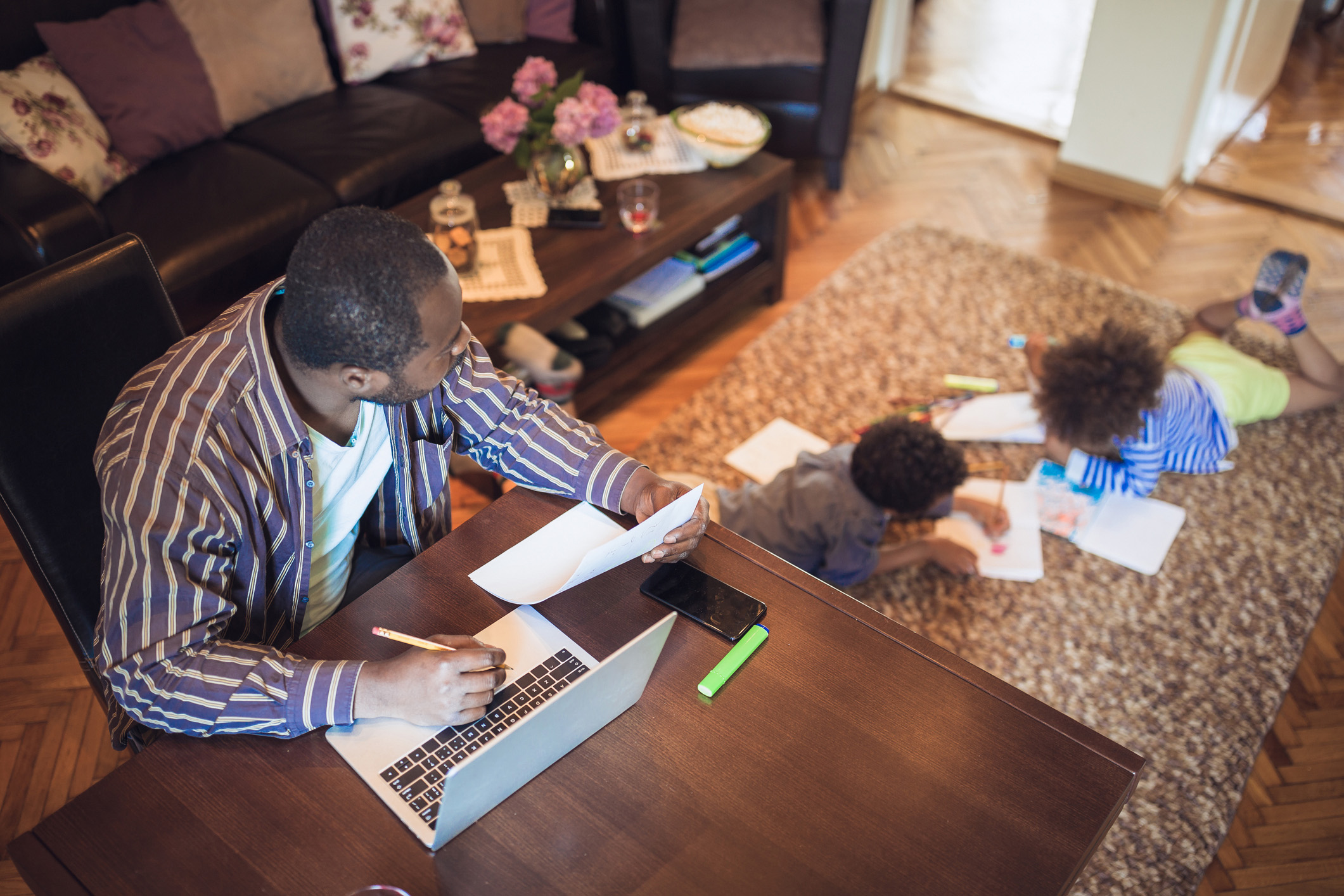 A father works on a class assignment from home on a laptop while his children draw on paper next to him.