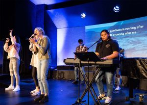 Four students playing piano, guitar and singing lead worship on a small stage