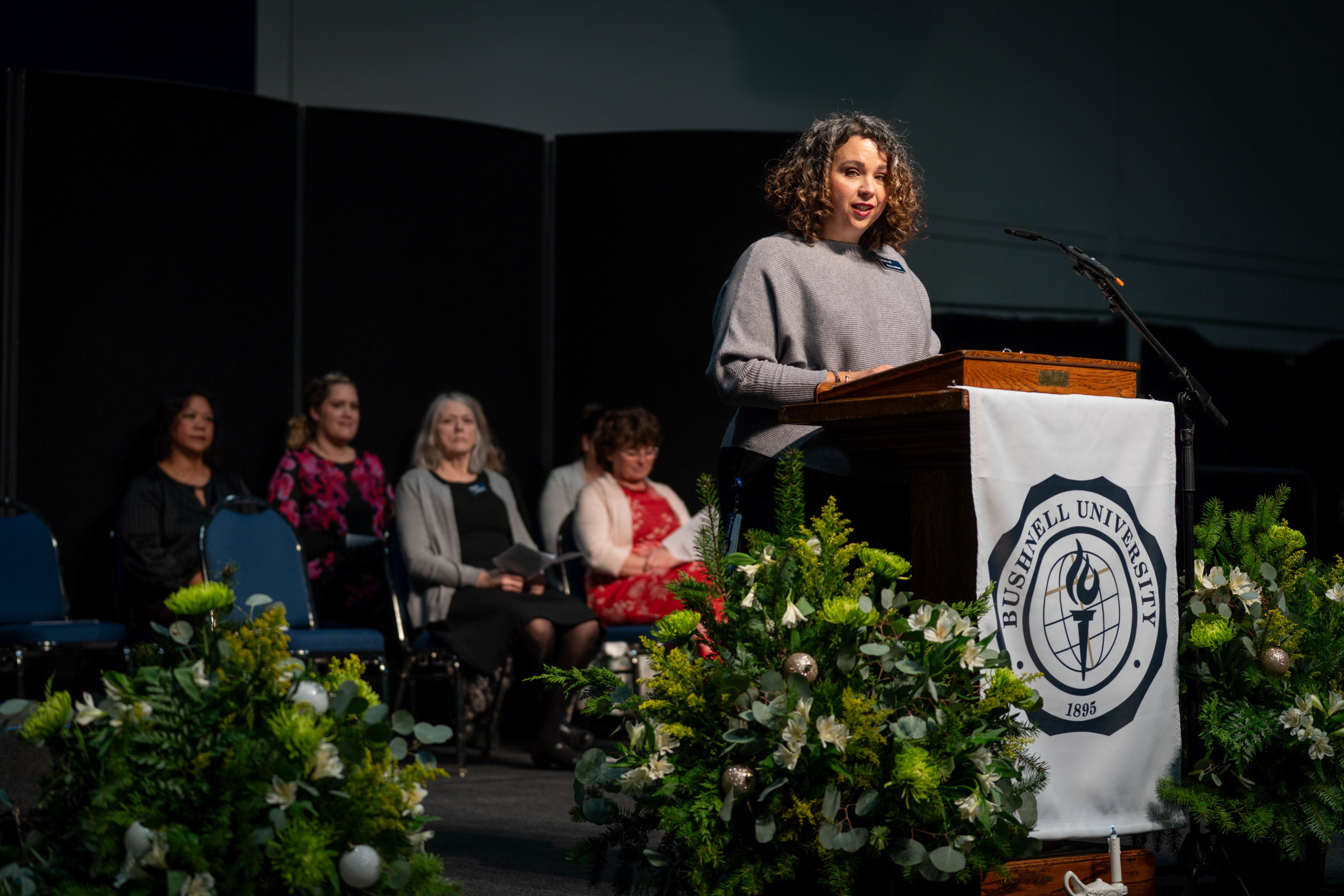 Bushnell University nursing professor Katy Lemieux speaks at a campus ceremony from a podium decorated with floral arrangements.