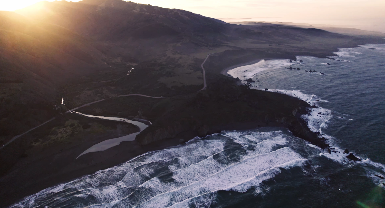 Aerial view of the Oregon Coast at sunrise with rugged cliffs, winding roads, and waves along the Pacific Ocean shoreline.