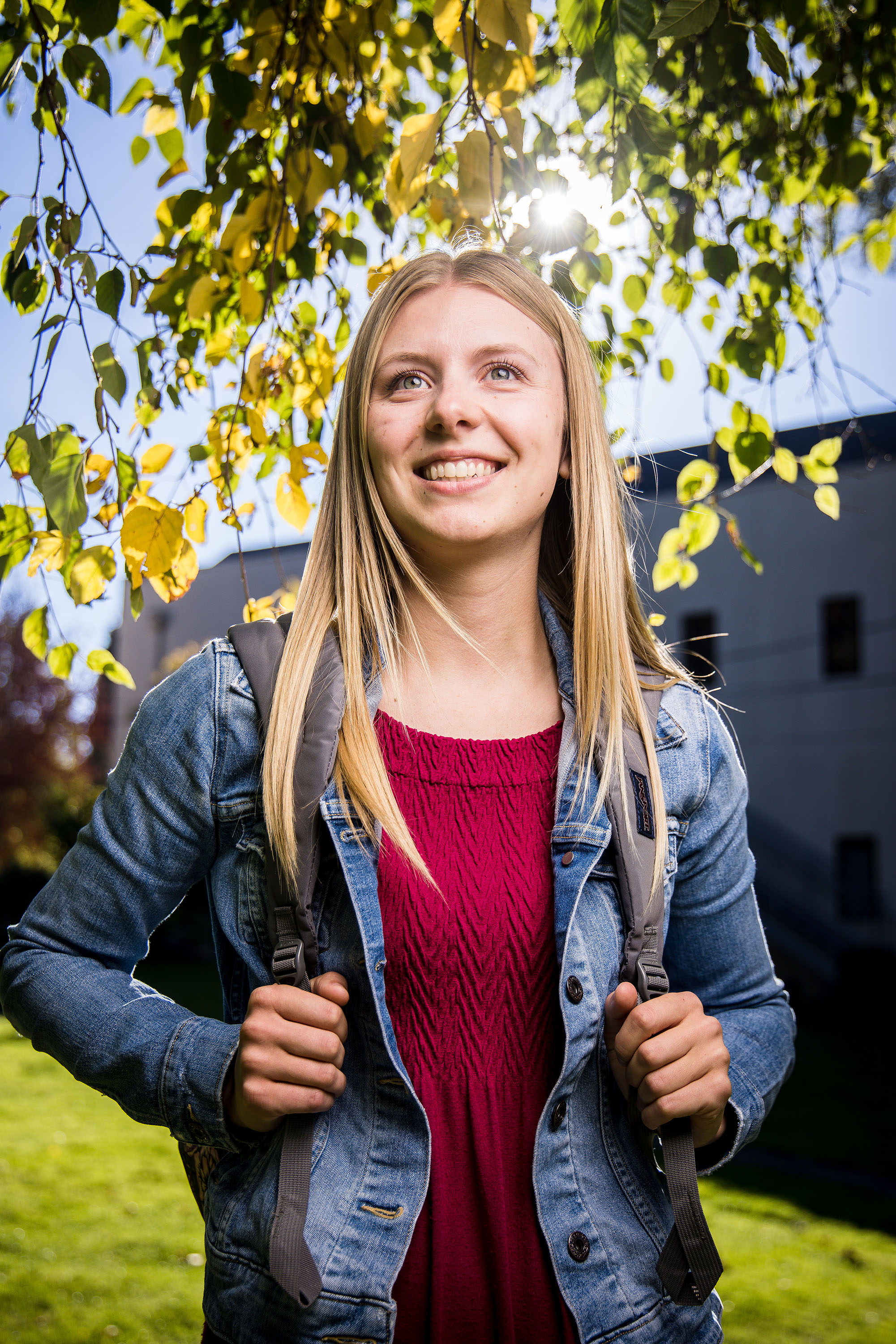 Student with long blonde hair and backpack smiling underneath a branch of green leaves with the sun behind.