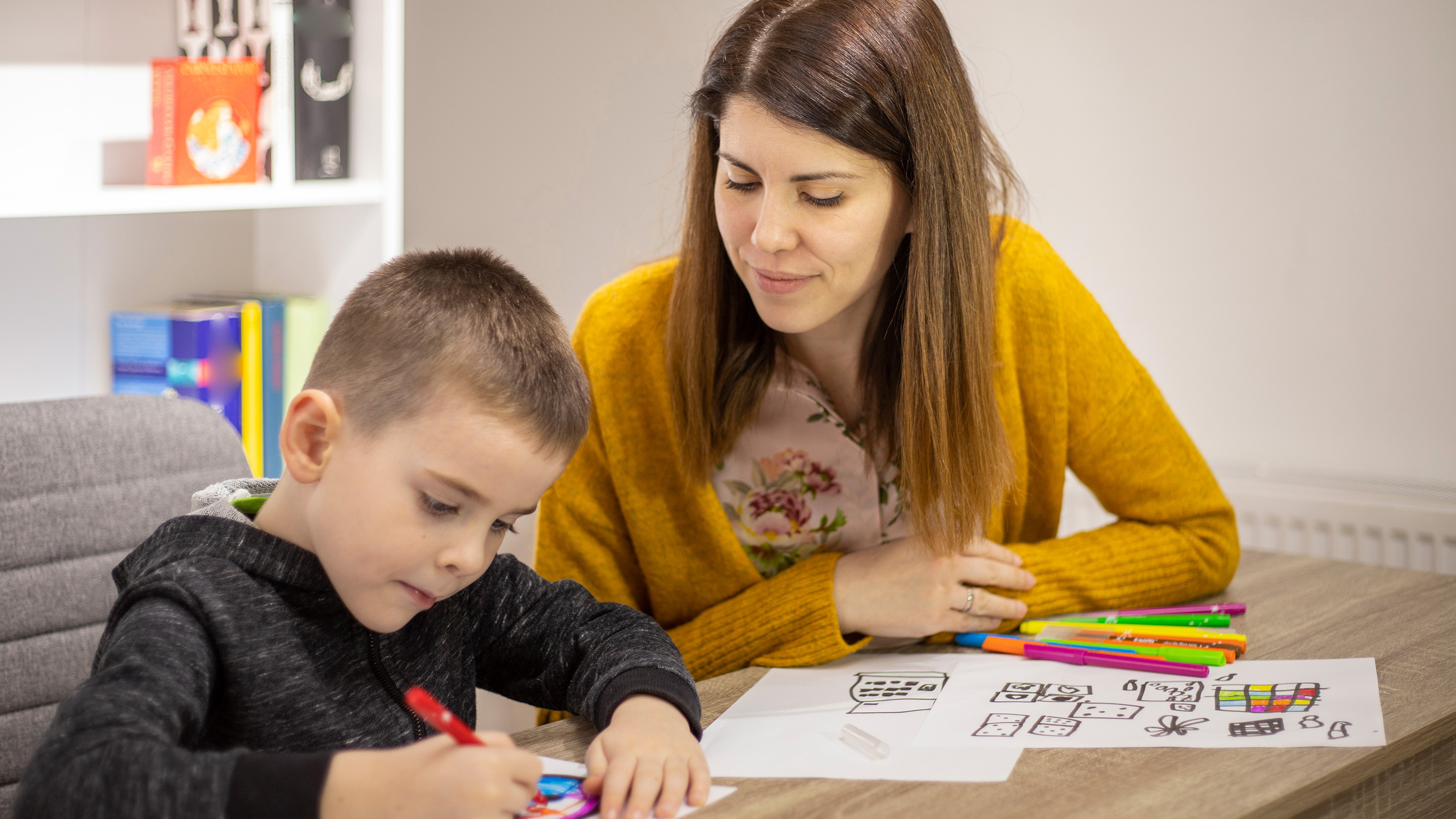 A school counselor works alongside a child as they draw pictures at a desk.