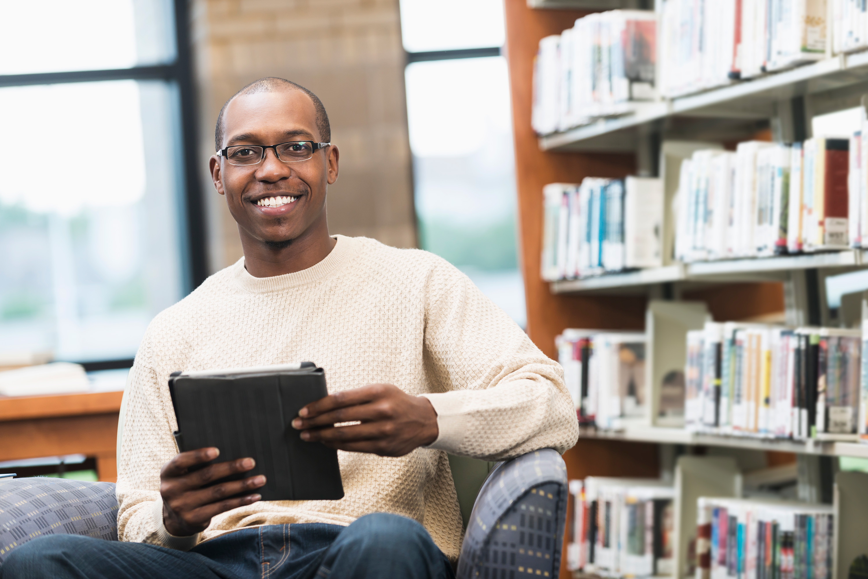 Student with glasses smiling while holding a tablet in a library setting.