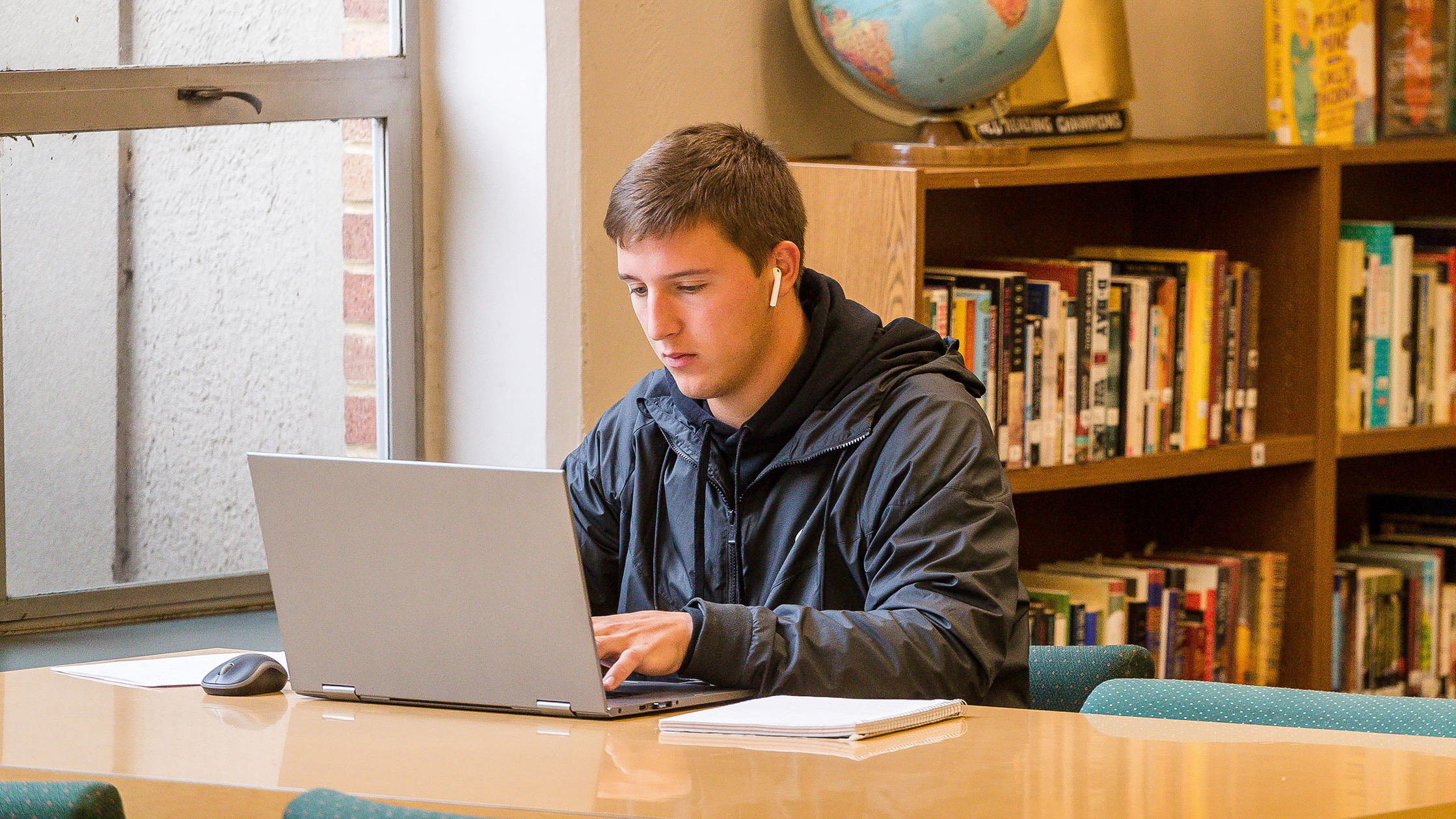 A student with earbuds focusses and types on their laptop in a library setting.