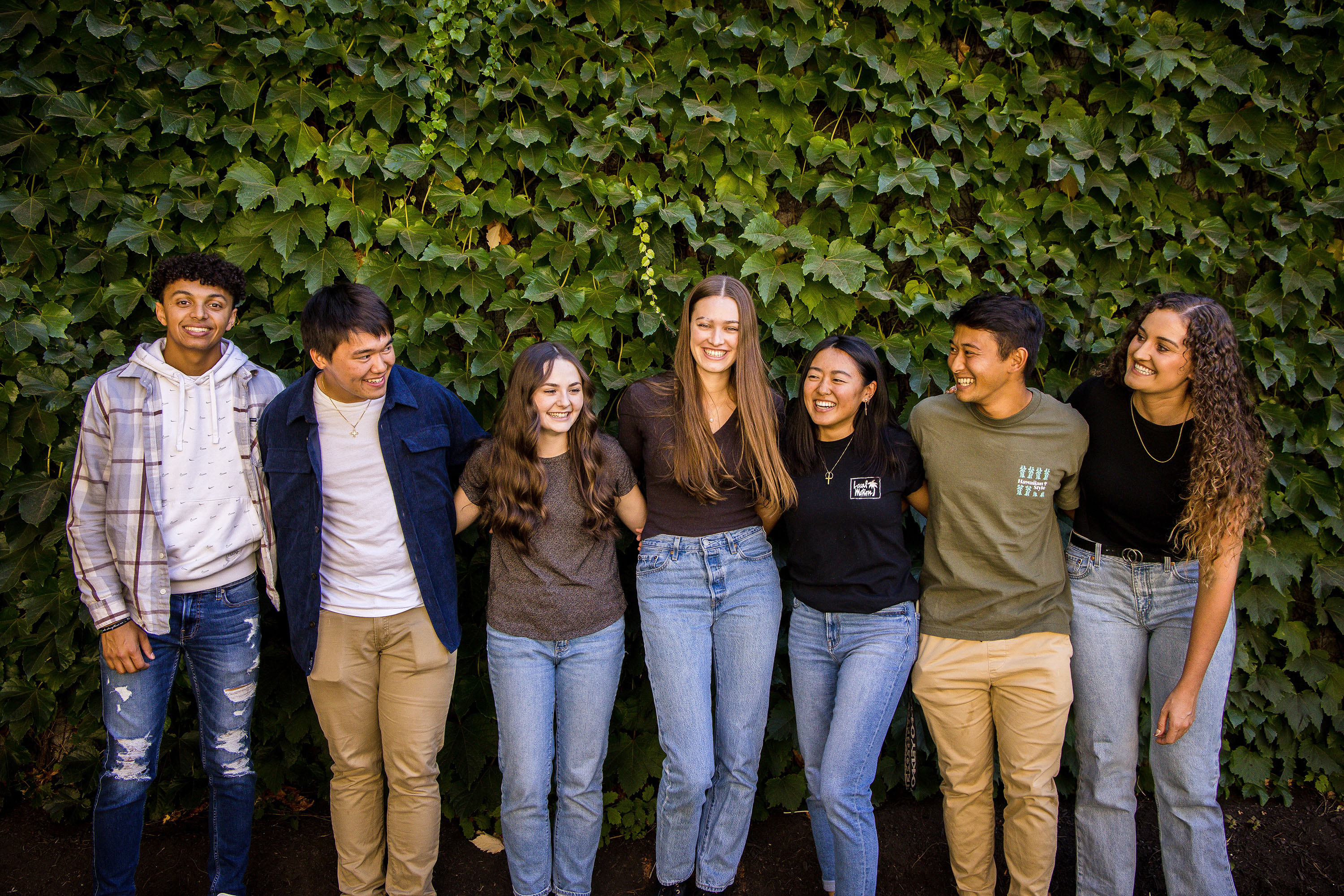 A group of students stand in a line with arms around each other laughing and smiling against a green ivy background.