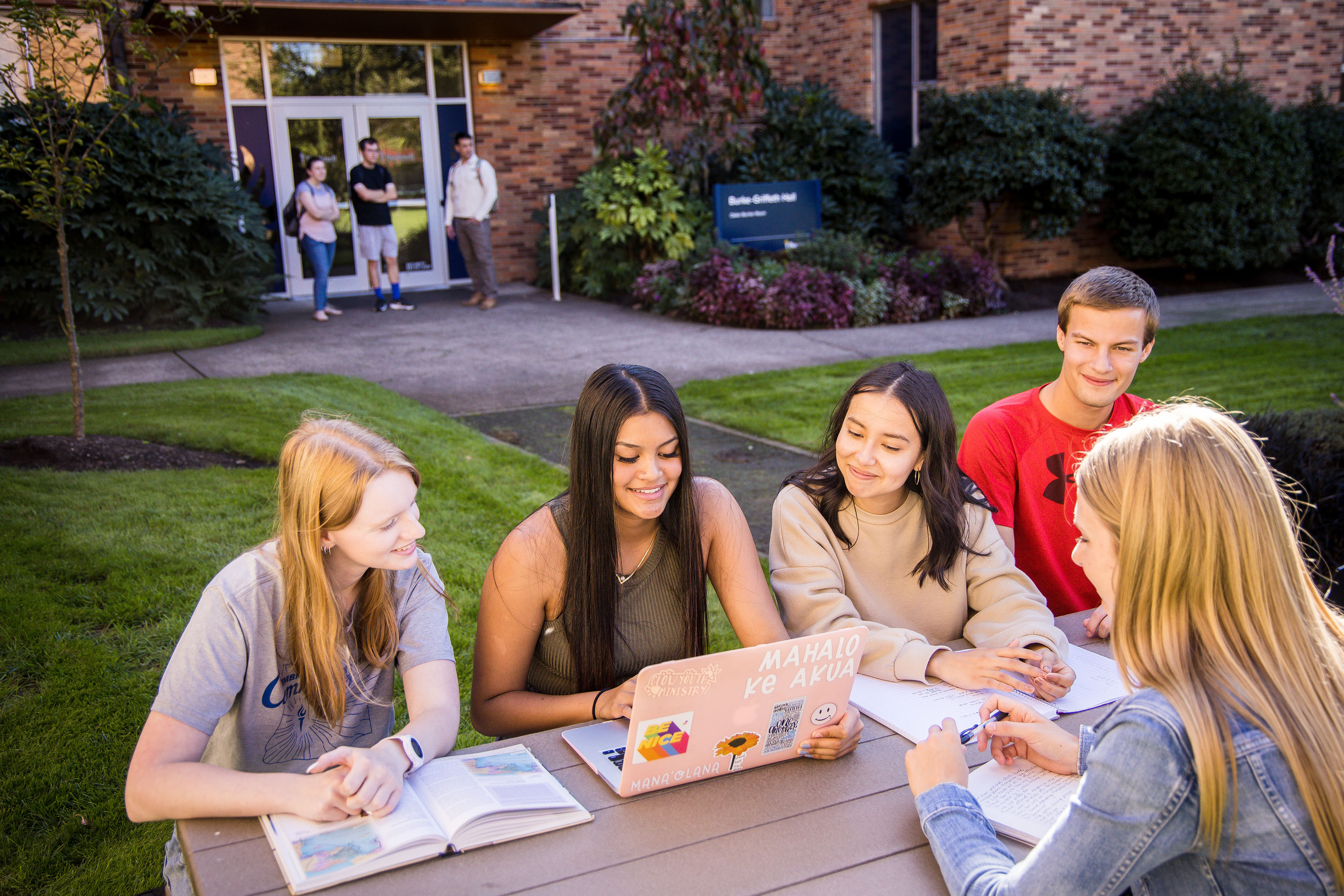 A group of students at an outdoor picnic table look together at a fellow student's laptop.
