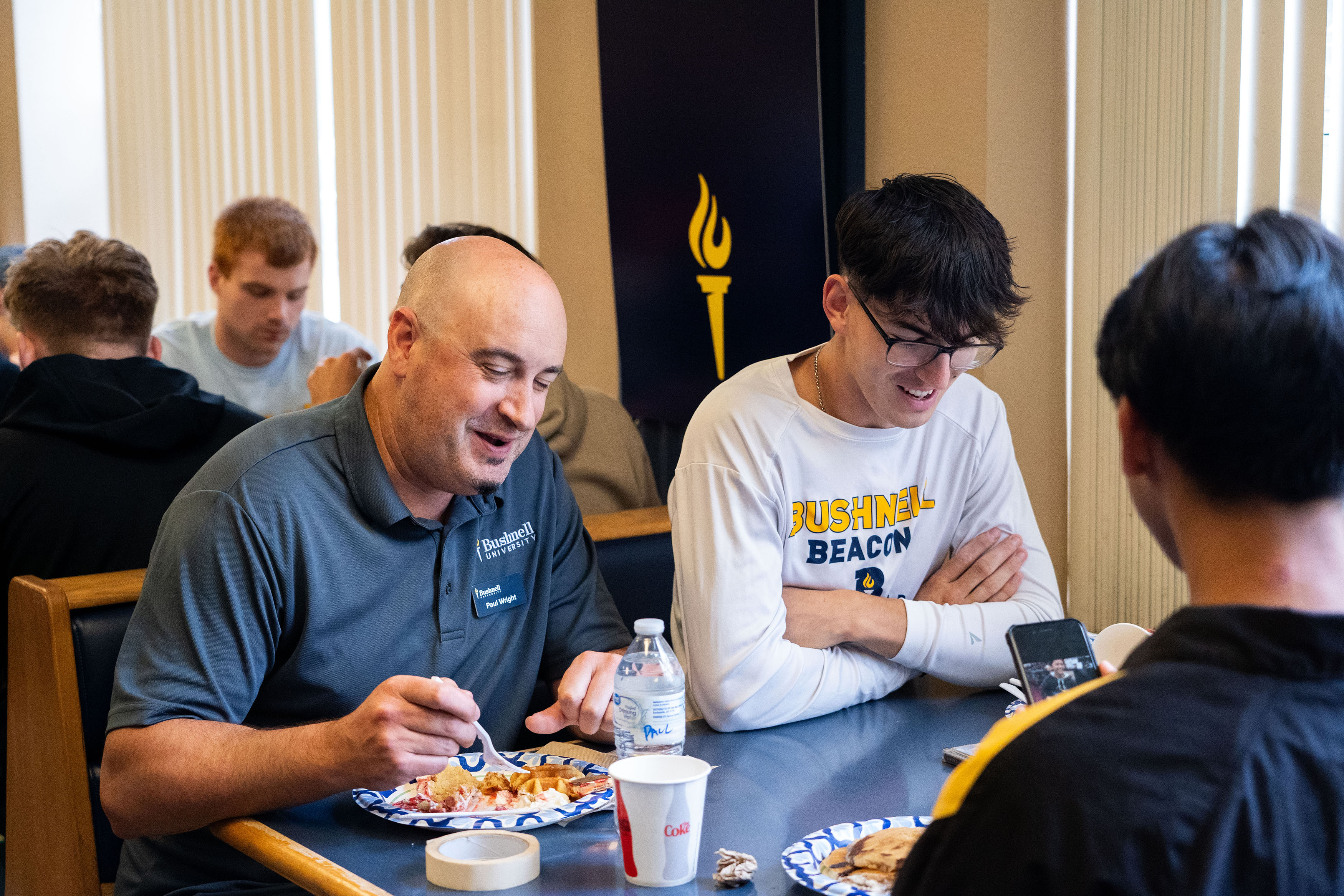 A Bushnell staff member and student laugh in a cafeteria booth next to a window while eating campus food.