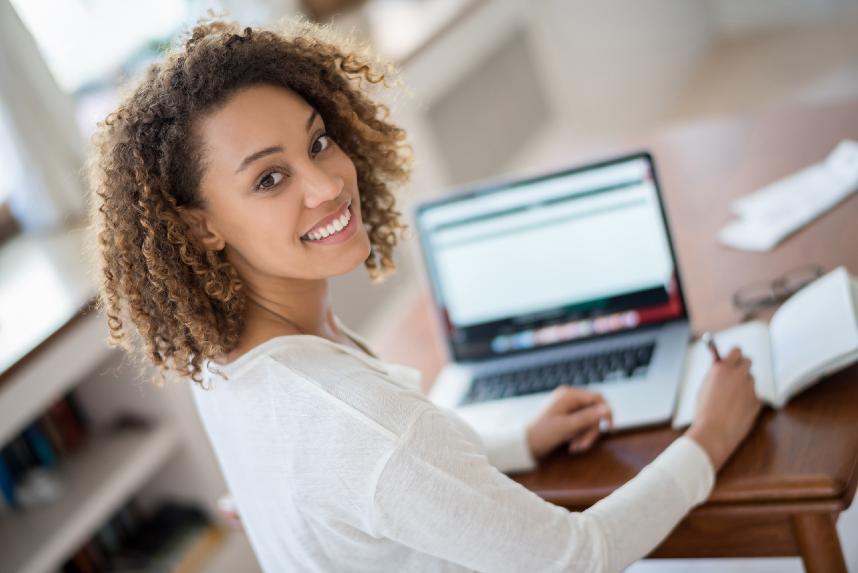 A student looks over her shoulder smiling at camera while studying her computer and writing in a notebook