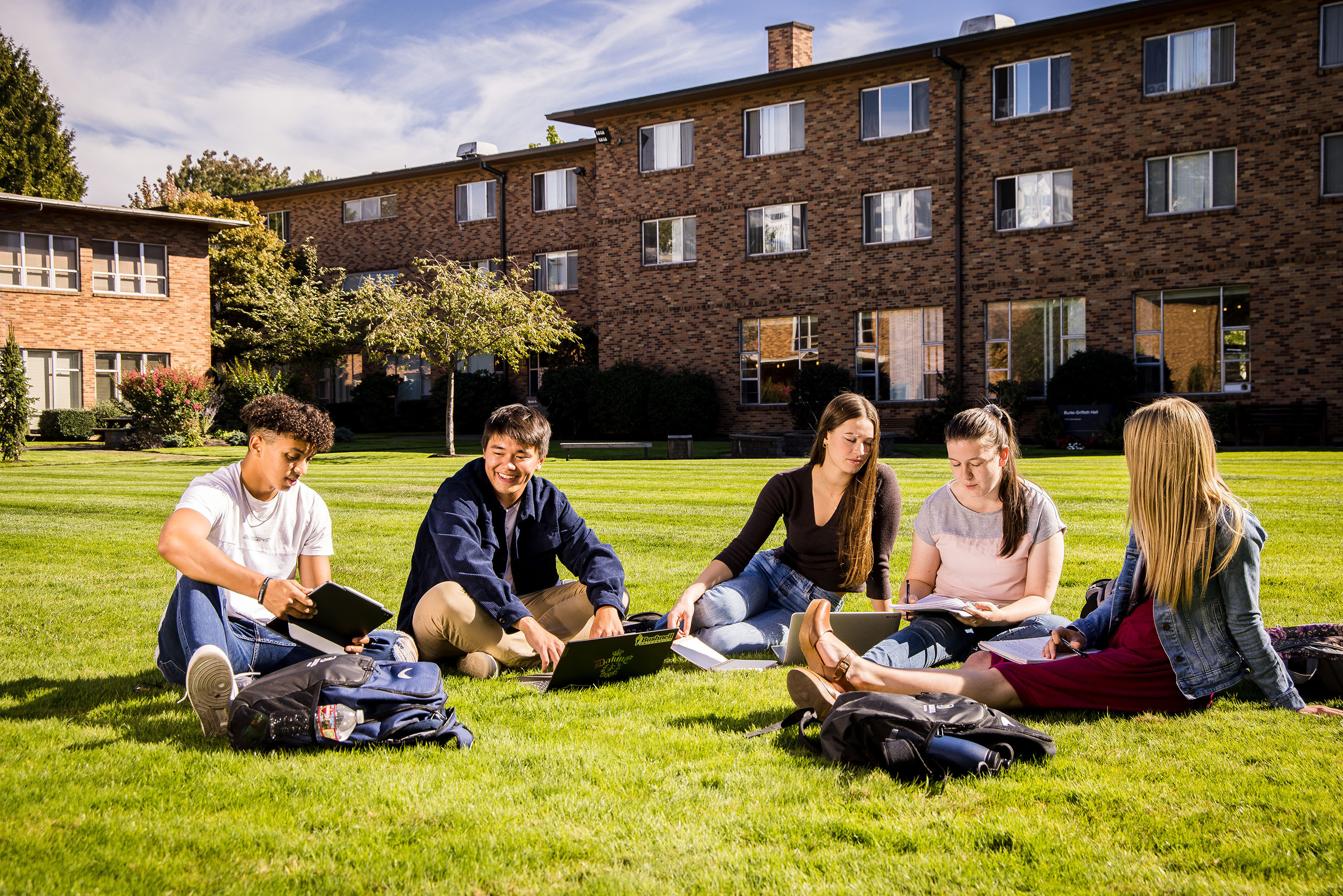 Five students sit with laptops, books, and homework discussing class in the grass of the Bushnell University Quad.